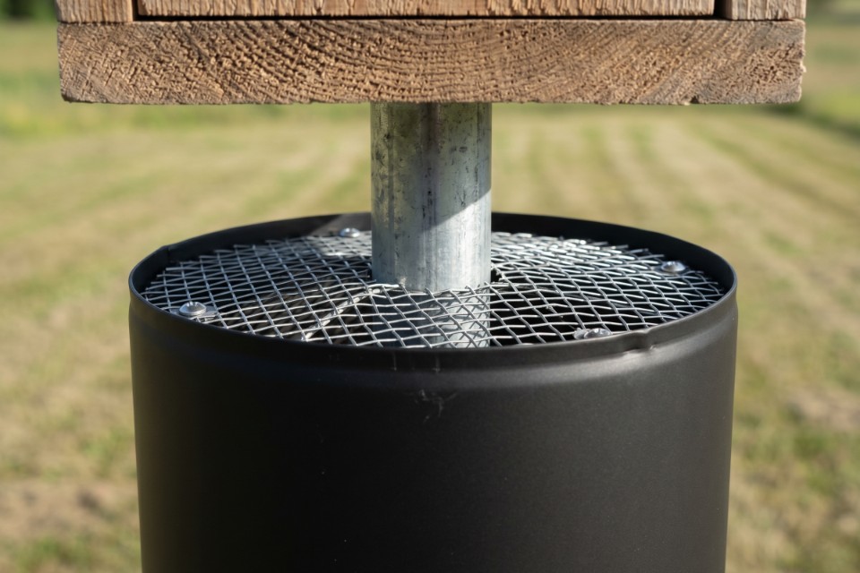 A forensic close-up of a stovepipe predator baffle showing the internal hardware cloth cap and centered galvanized mounting pole for a bluebird house.
