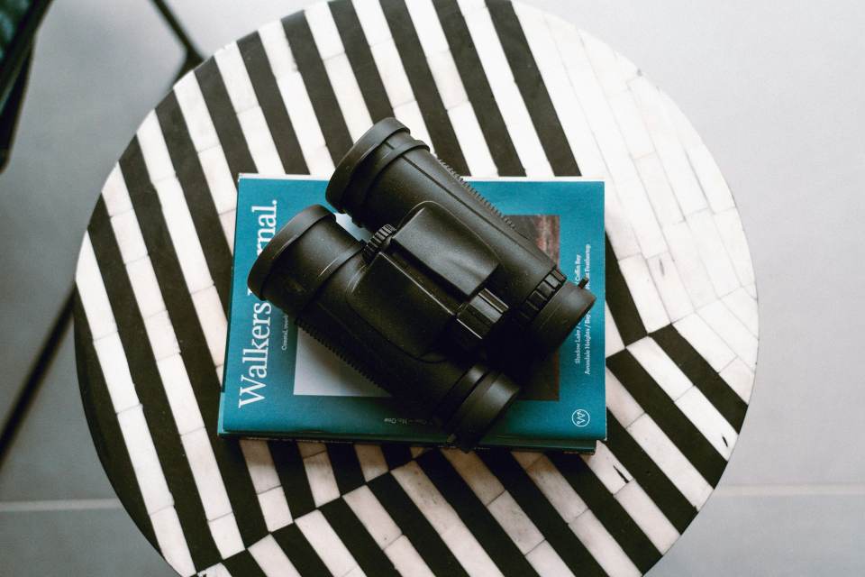 A black roof-prism birding binocular resting on a teal field notebook on a black and white striped round table.