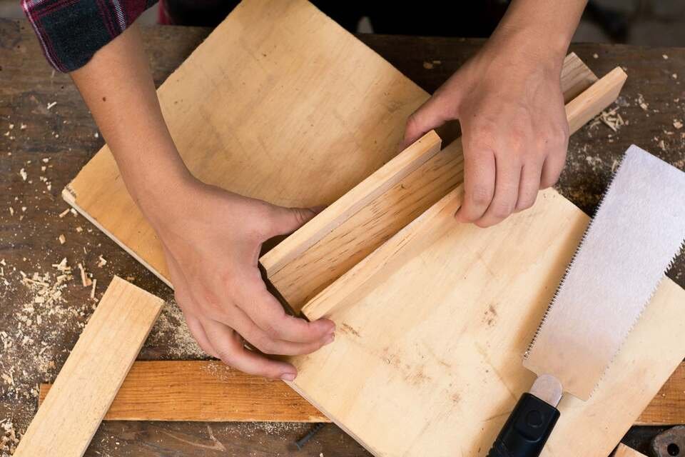 Close-up of a person's hands using a hand saw and wooden boards on a workshop bench covered in sawdust.